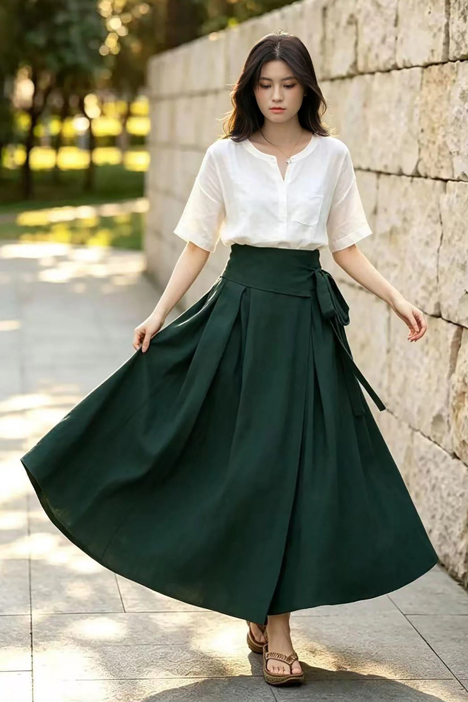 May include: A white short-sleeved blouse with a V-neckline and a single chest pocket paired with a high-waisted dark forest green wrap skirt. The skirt features a wide waistband with a side tie closure and deep pleats that create a full, flowing silhouette.