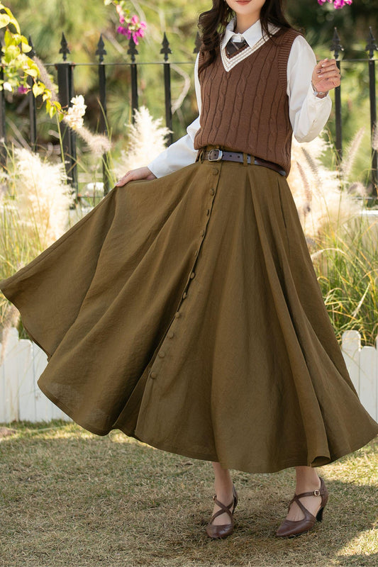 May include: An olive green button-down skirt with a wide, flowing silhouette. The skirt is paired with a brown cable-knit sweater vest, a white collared shirt, and a brown belt. The outfit is completed with brown heels. The background features a white picket fence and greenery.