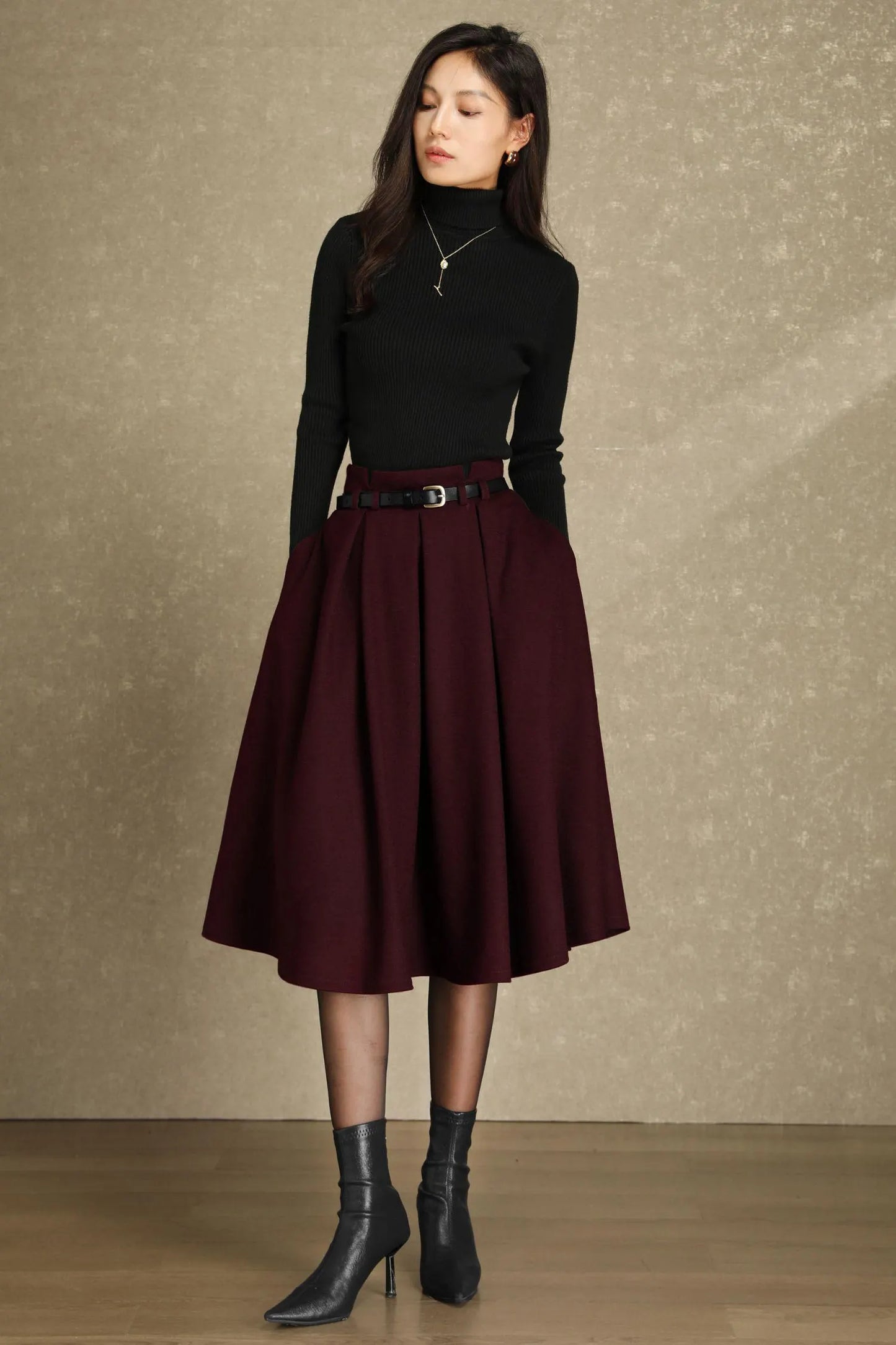 May include: A woman wearing a black turtleneck sweater, a burgundy pleated skirt, and black ankle boots. The skirt is cinched at the waist with a black belt. The outfit is suitable for fall or winter wear.
